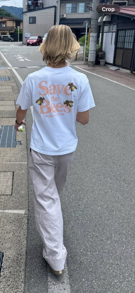 A person with blond hair walks away from the camera, wearing a white t-shirt that says "Save the Bees" with bee illustrations.
