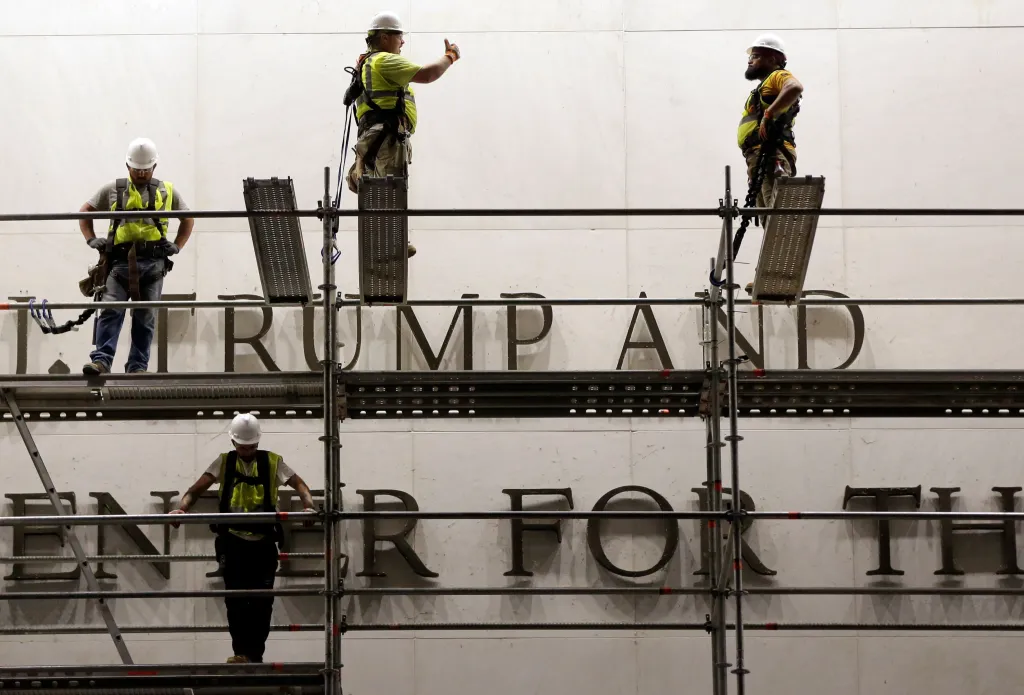 Construction workers on scaffolding in front of a white building with the letters "TRUMP AND" and "ENTER FOR TH" visible.