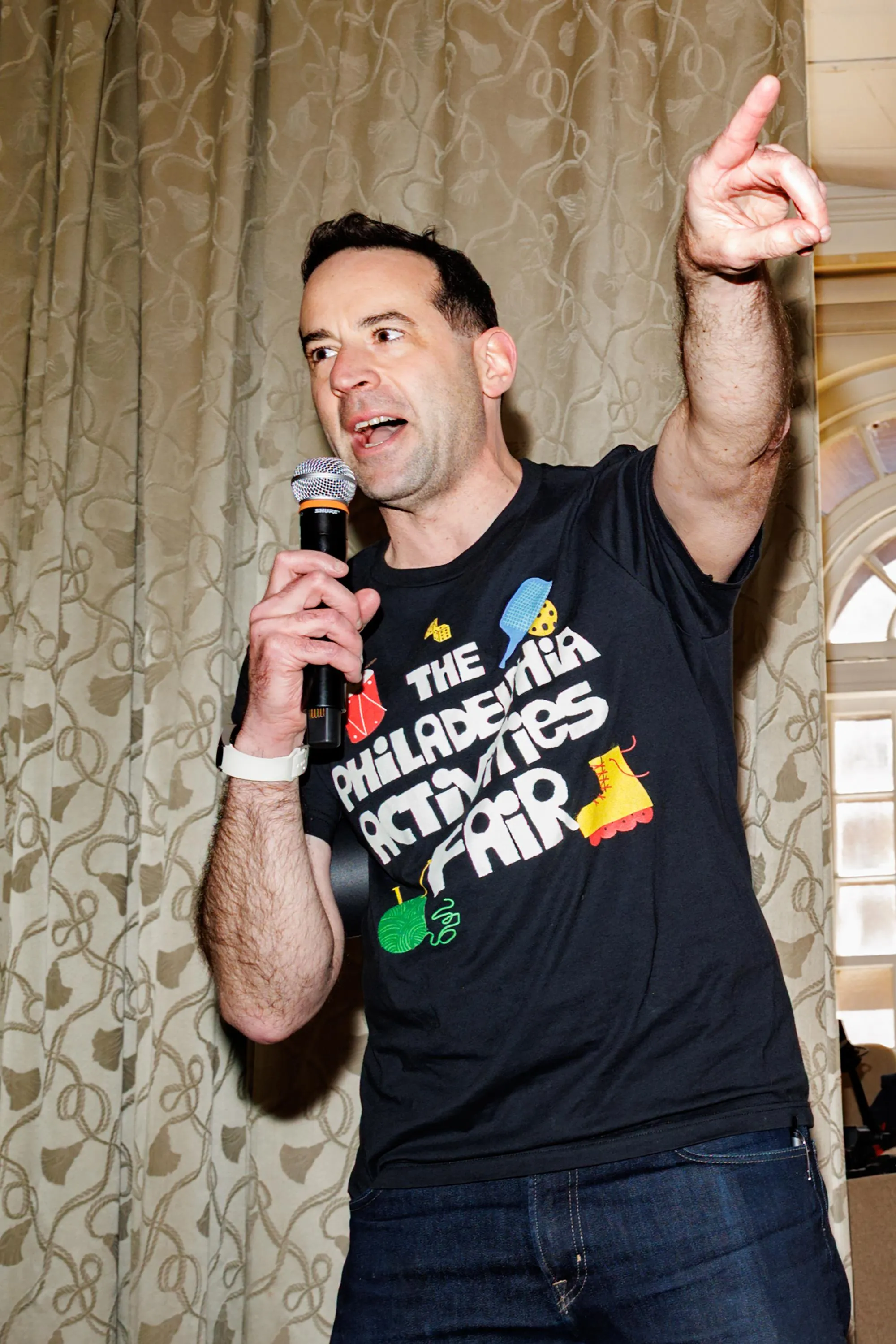 a man animatedly speaks into a microphone while pointing out of frame. His shirt reads “The Philadelphia Activities Fair” a man animatedly speaks into a microphone while pointing out of frame. His shirt reads “The Philadelphia Activities Fair”