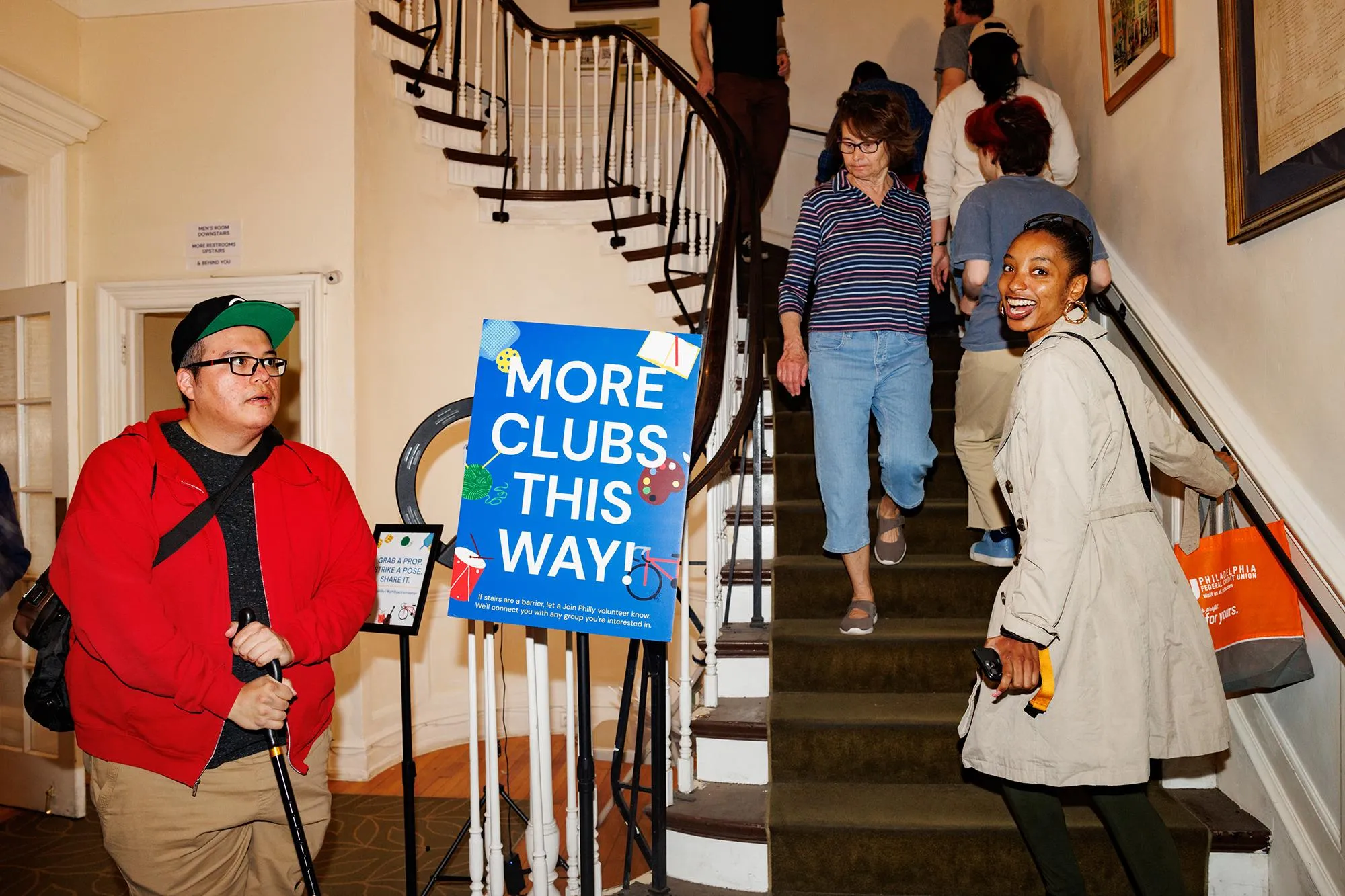 a woman smiling at the camera as she ascents a staircase full of people. A nearby blue sign reads “More clubs this way!” a woman smiling at the camera as she ascents a staircase full of people. A nearby blue sign reads “More clubs this way!”