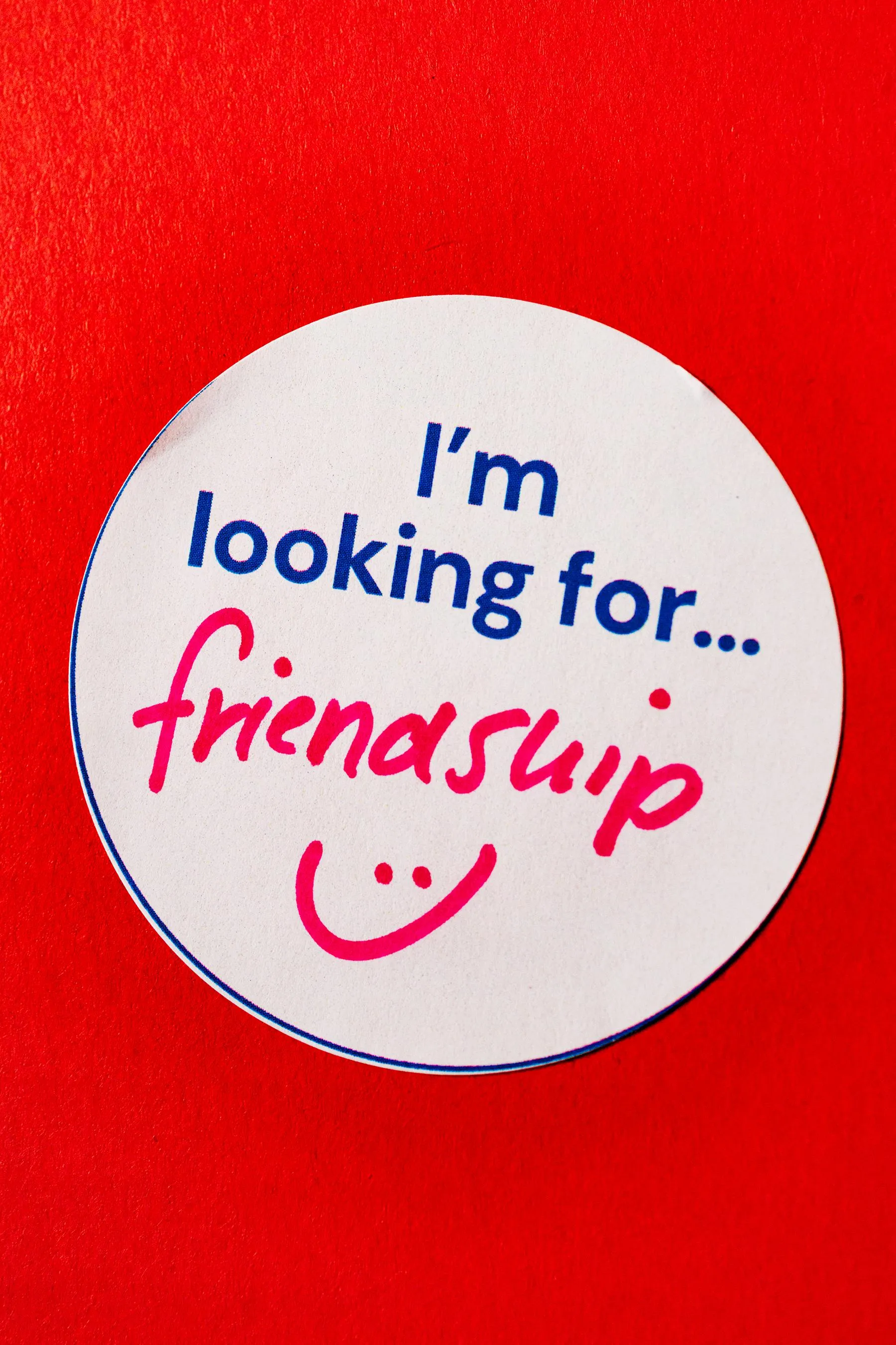 a sticker reading “I’m looking for… friendship” with a hand-drawn smiley face is stuck to a red wall a sticker reading “I’m looking for… friendship” with a hand-drawn smiley face is stuck to a red wall