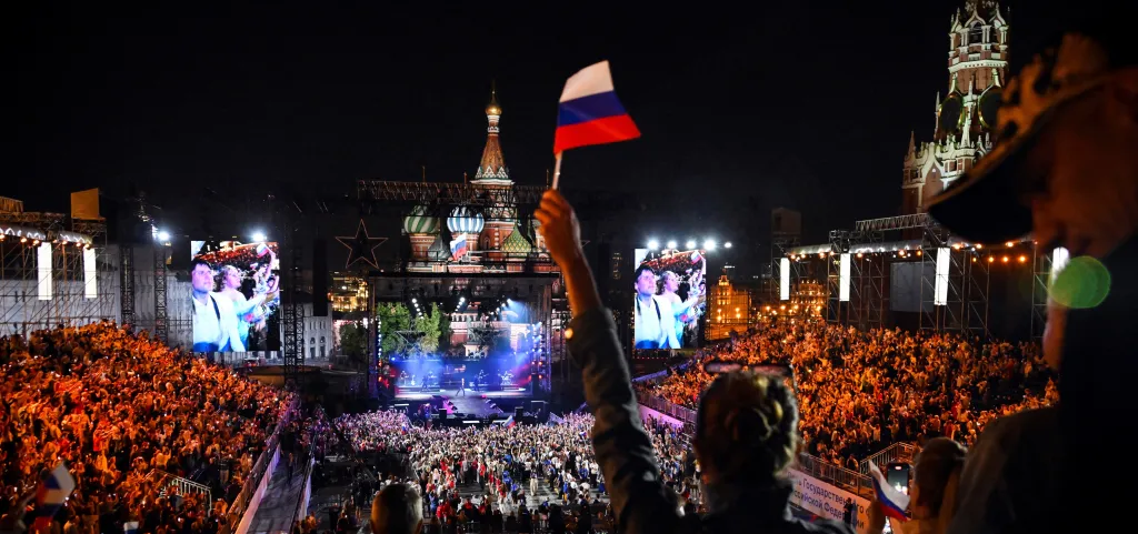 A large crowd of people gathered at night, with a person holding a Russian flag in the foreground, and a stage with a performance and large screens in the background.