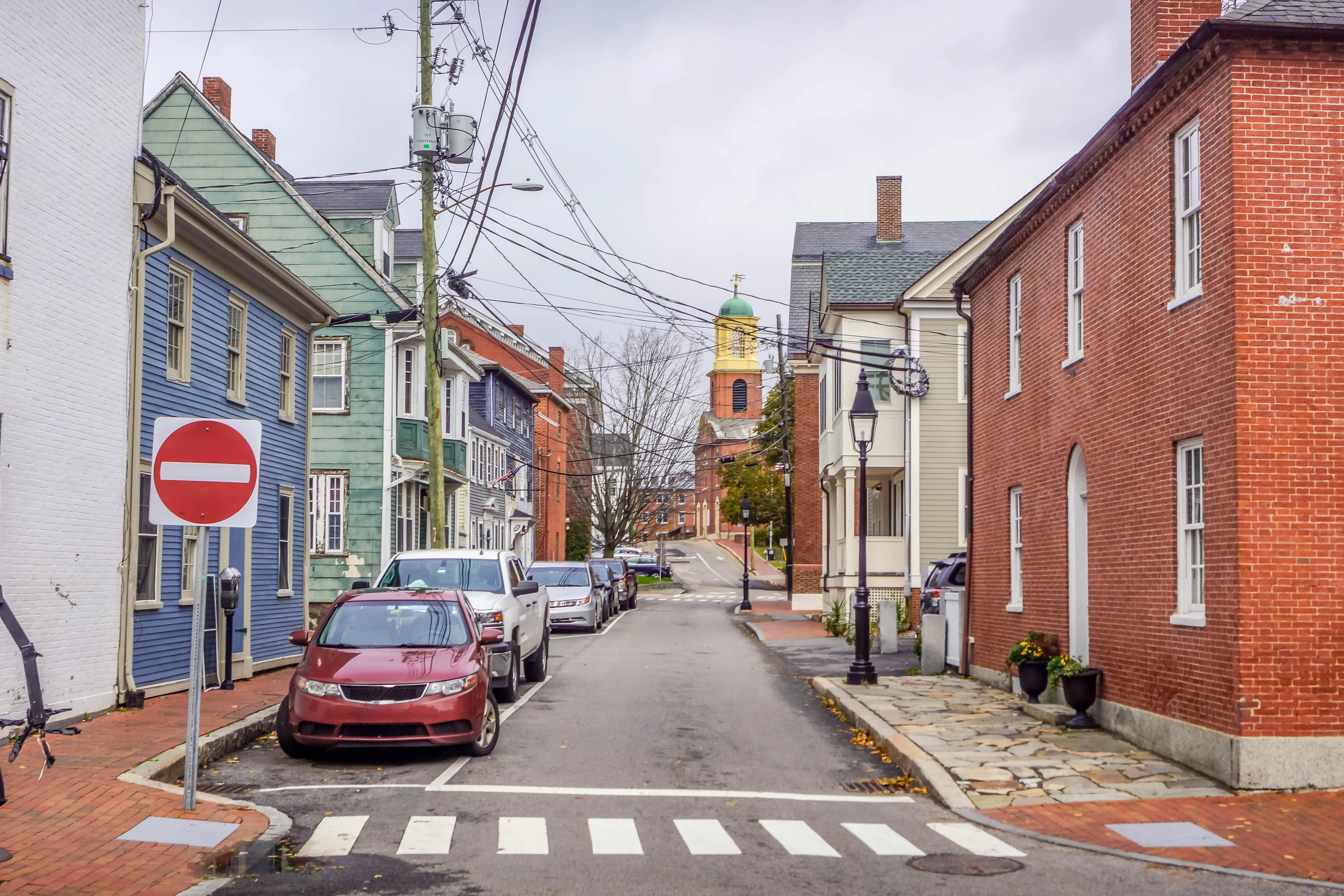 A narrow street in an older, built-up neighborhood lined with historic brick and clapboard buildings, parked cars, brick sidewalks, overhead utility wires, and a church tower in the background.