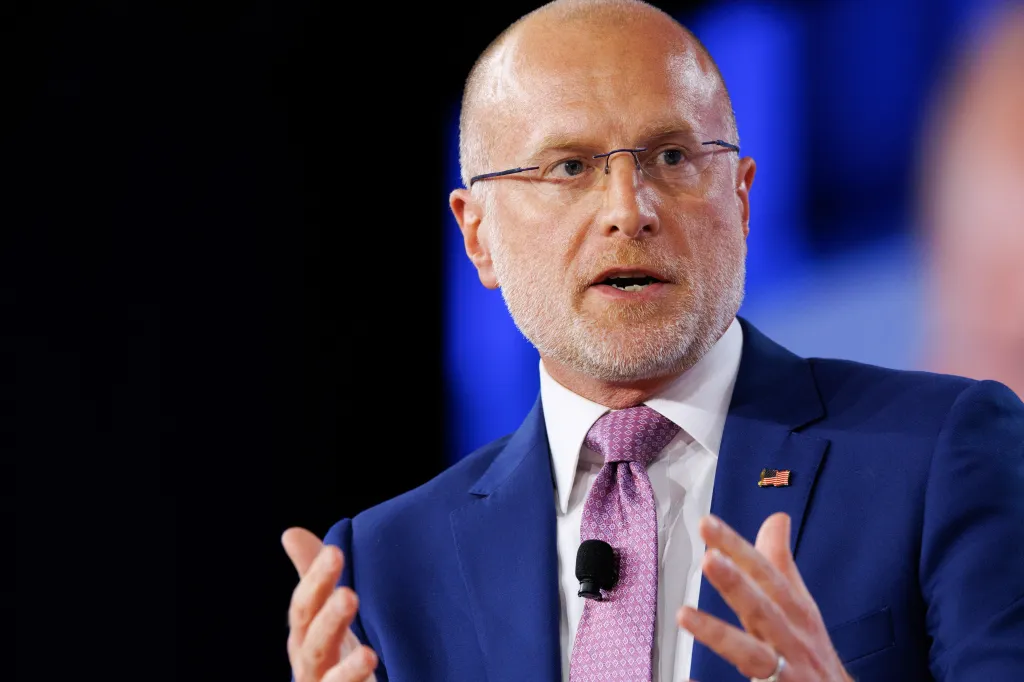A man with a bald head, glasses, and a beard speaks into a microphone clipped to his purple tie, wearing a blue suit jacket with an American flag pin.