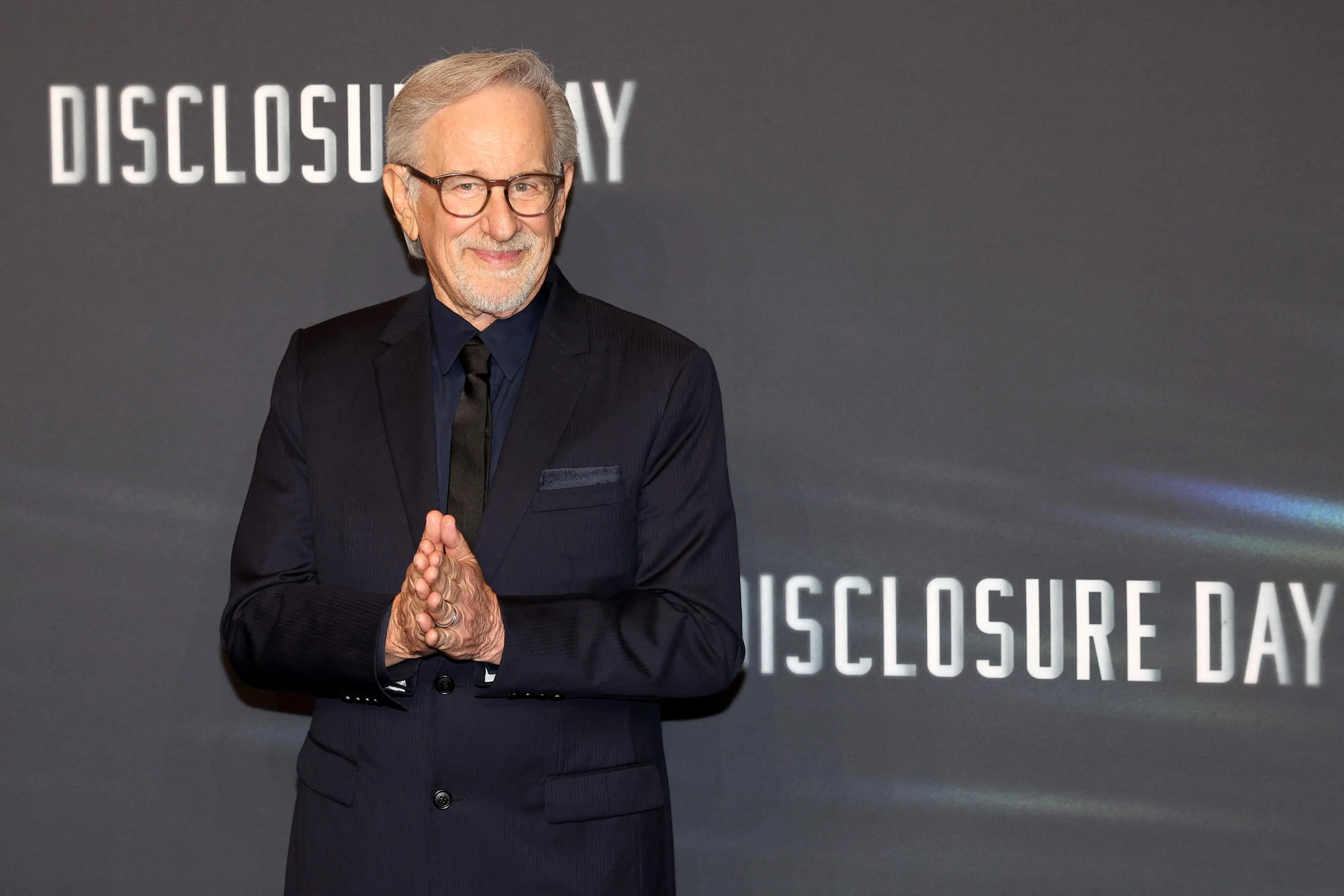 Steven Spielberg, in a black suit, stands in front of a backdrop reading “Disclosure Day.”