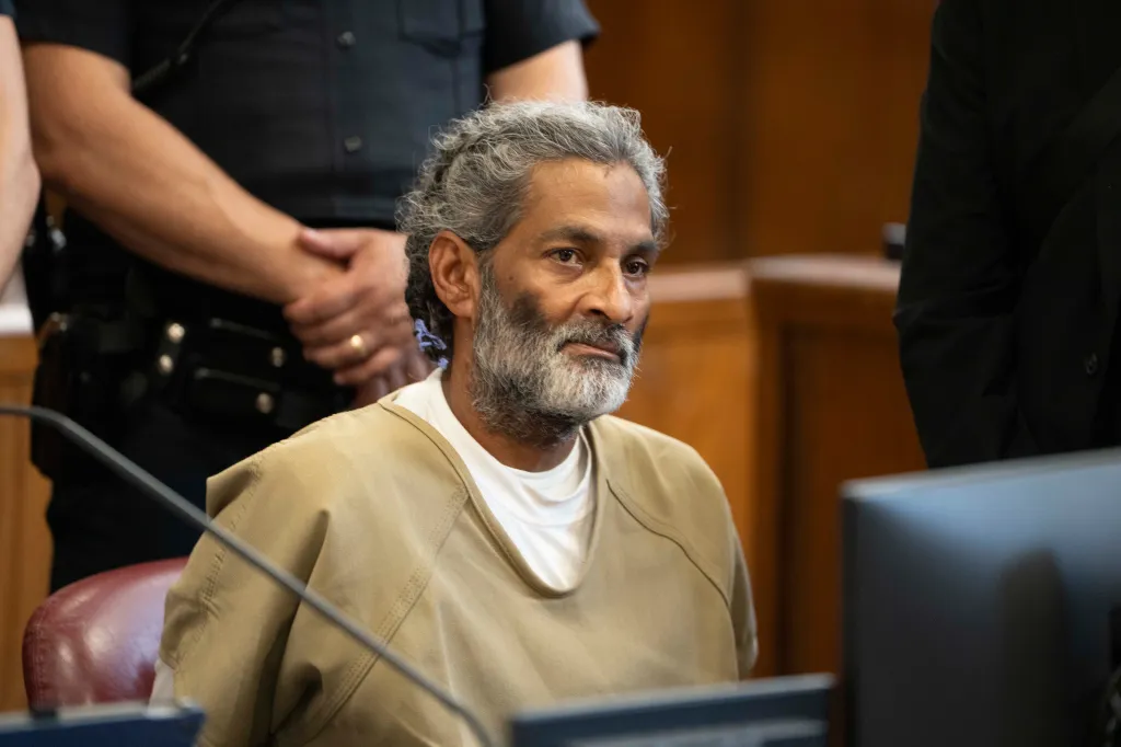 Hector Deleon in court, wearing an tan jumpsuit over a white undershirt, with a gray beard and gray curly hair.
