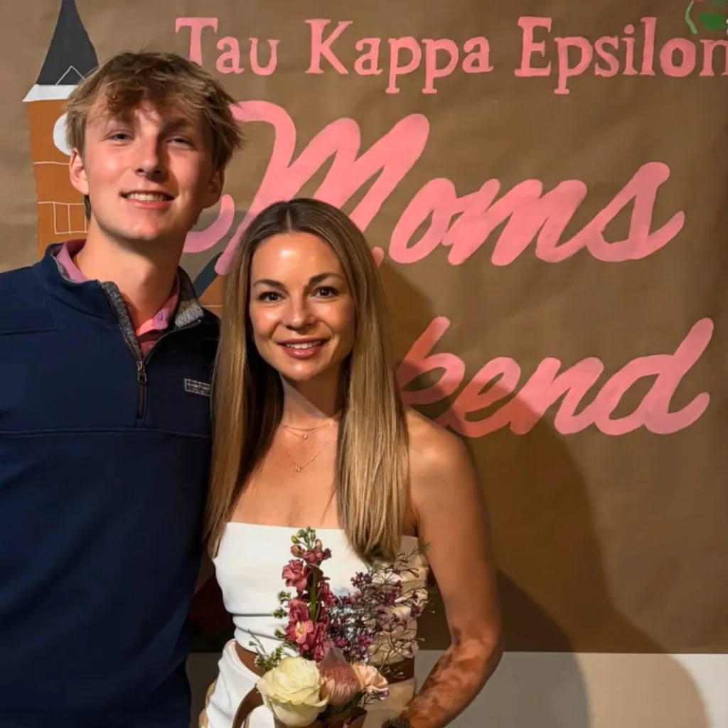 A man and a woman stand in front of a banner that reads "Tau Kappa Epsilon Moms Weekend."