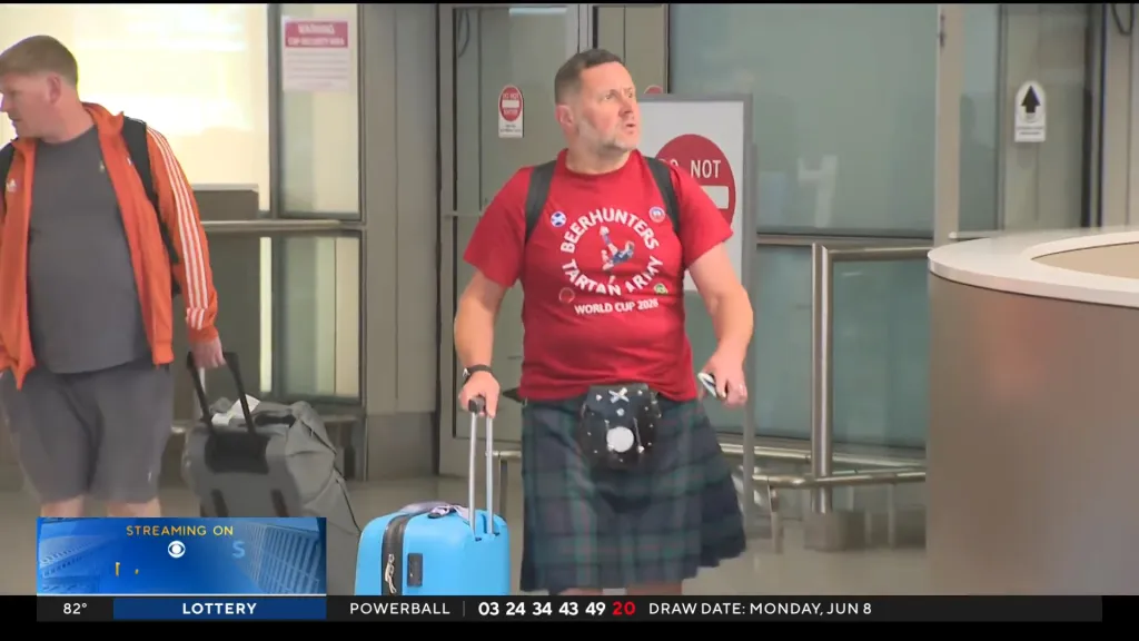 A man in a red "Beerhunters Tartan Army World Cup 2026" t-shirt and kilt pulling a light blue suitcase through an airport.