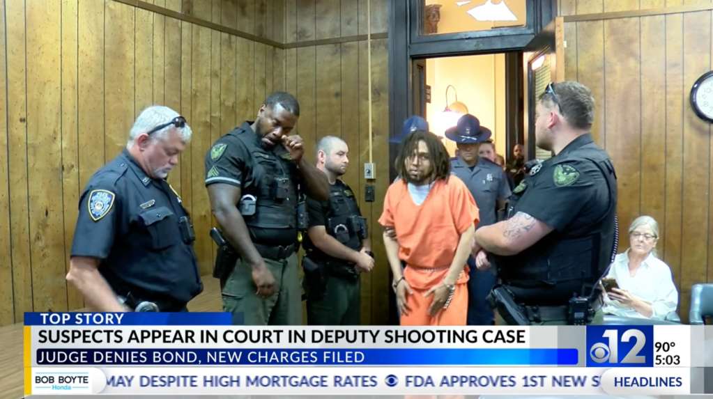A man in an orange jumpsuit is escorted by officers in a courtroom, with text "SUSPECTS APPEAR IN COURT IN DEPUTY SHOOTING CASE" and "JUDGE DENIES BOND, NEW CHARGES FILED."