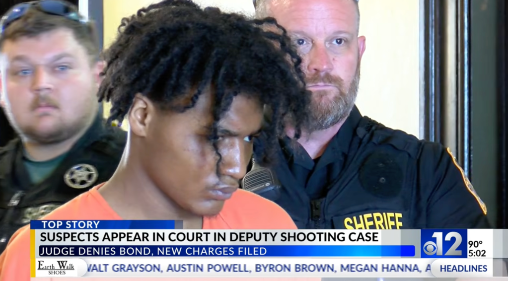 A man in an orange jumpsuit with dark dreadlocks appears in court, flanked by two sheriffs, as a news chyron reads, "Suspects appear in court in deputy shooting case, judge denies bond, new charges filed."