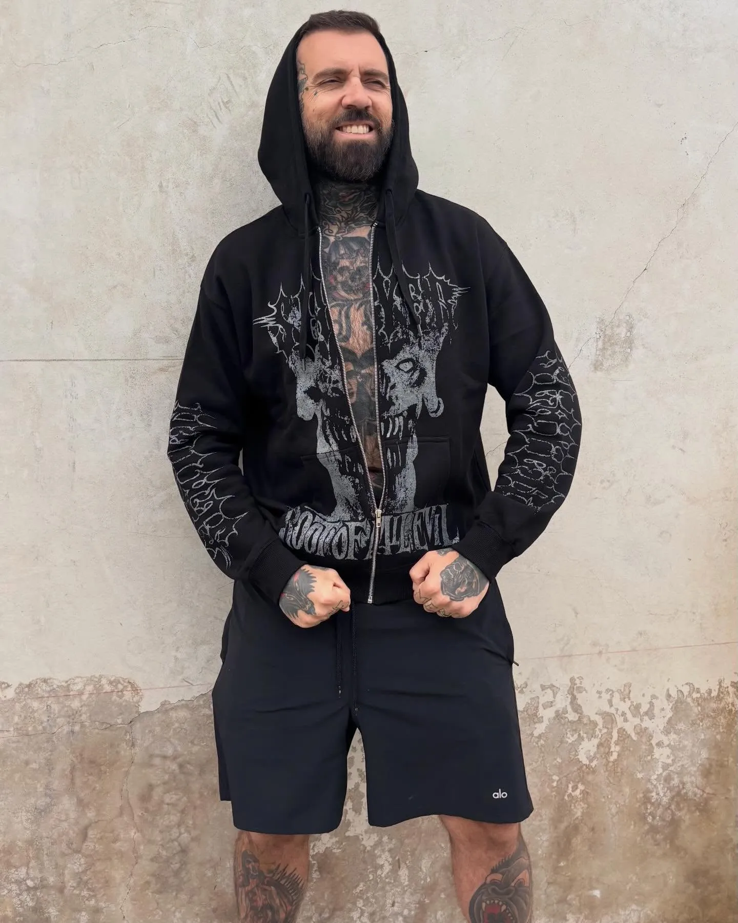 A man with a full beard and tattoos, wearing a black hooded sweatshirt with a skull design and black shorts, stands smiling against a textured wall.