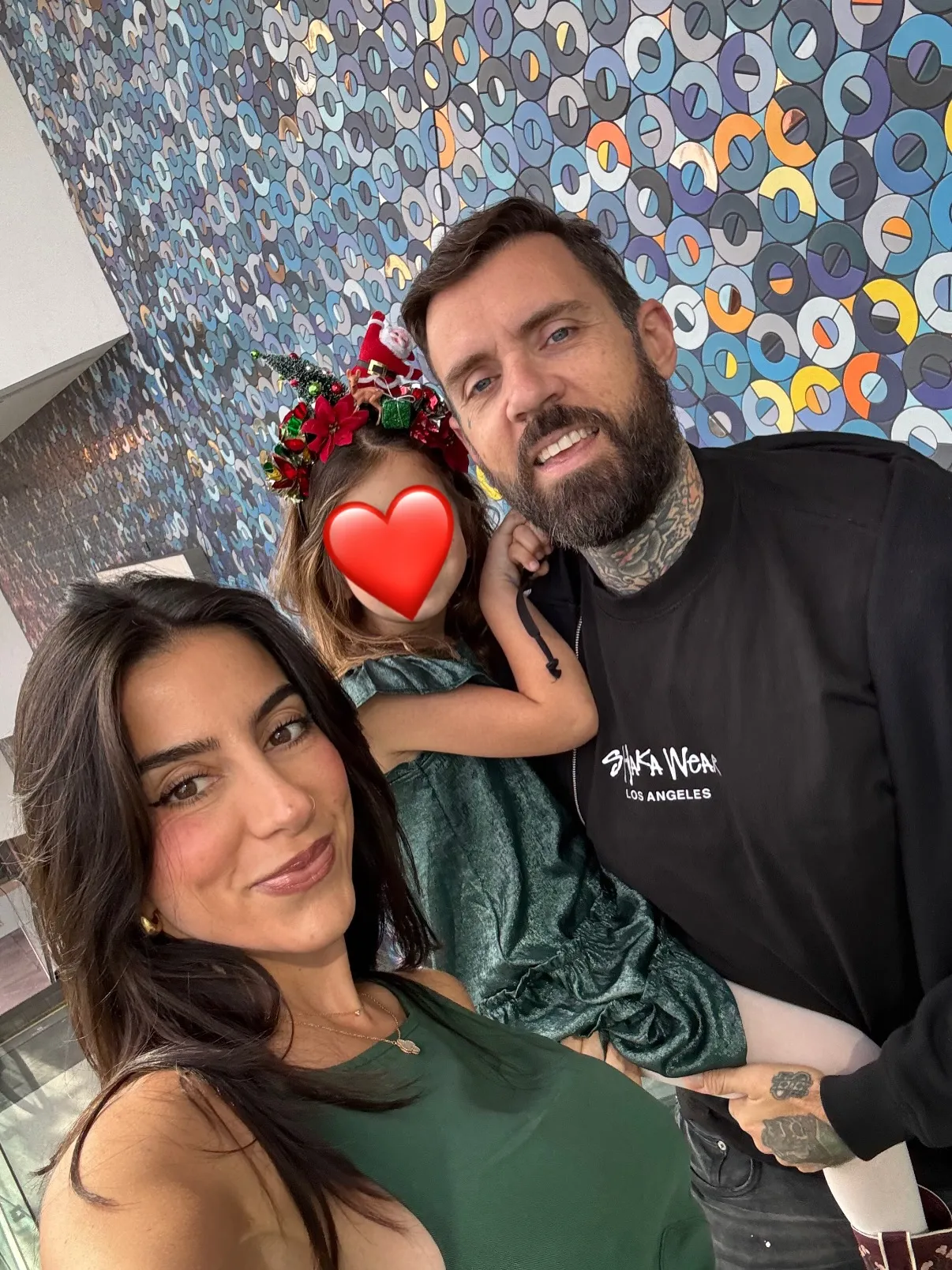 Adam22, Lena the Plug, and their child, whose face is covered by a red heart, pose for a selfie in front of a colorful, patterned wall.