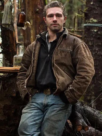 Matt Brown wearing a brown jacket and jeans, standing in a wooded area.