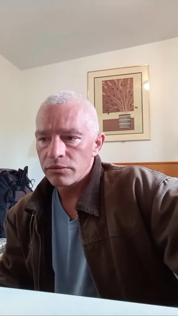A man with short gray hair and a brown jacket looks down with a concerned expression.