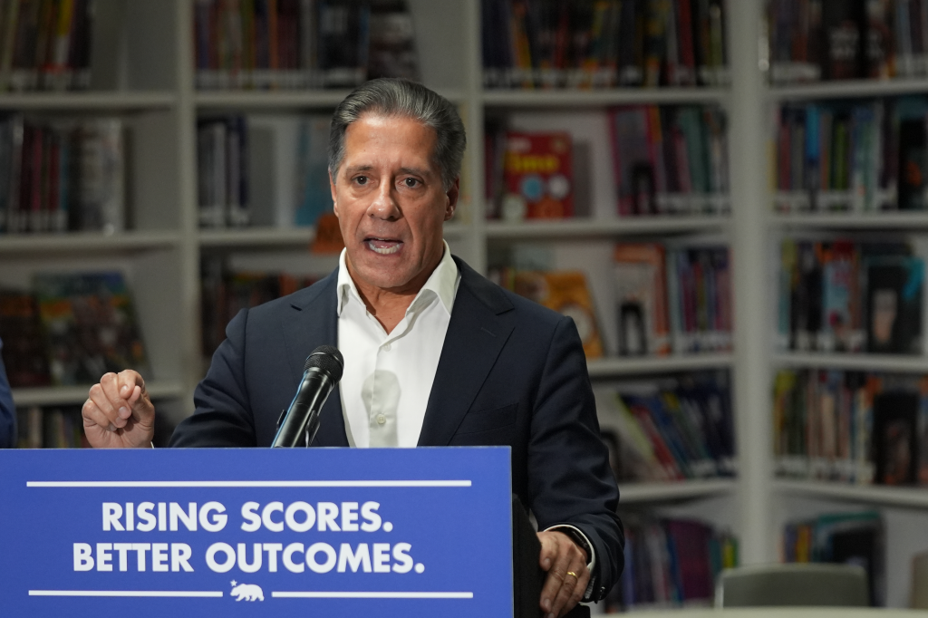 Superintendent Alberto Carvalho speaking at a podium with the text "RISING SCORES. BETTER OUTCOMES." in front of a bookshelf.