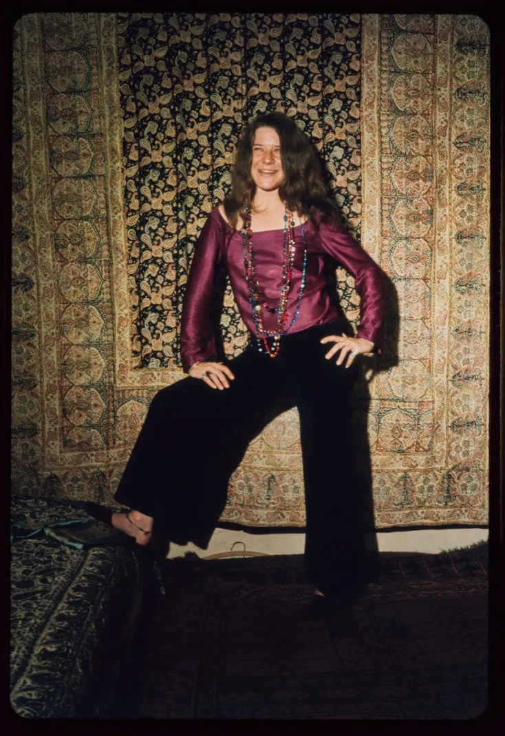 Janis Joplin in a vibrant purple top and black wide-leg pants against a paisley background.