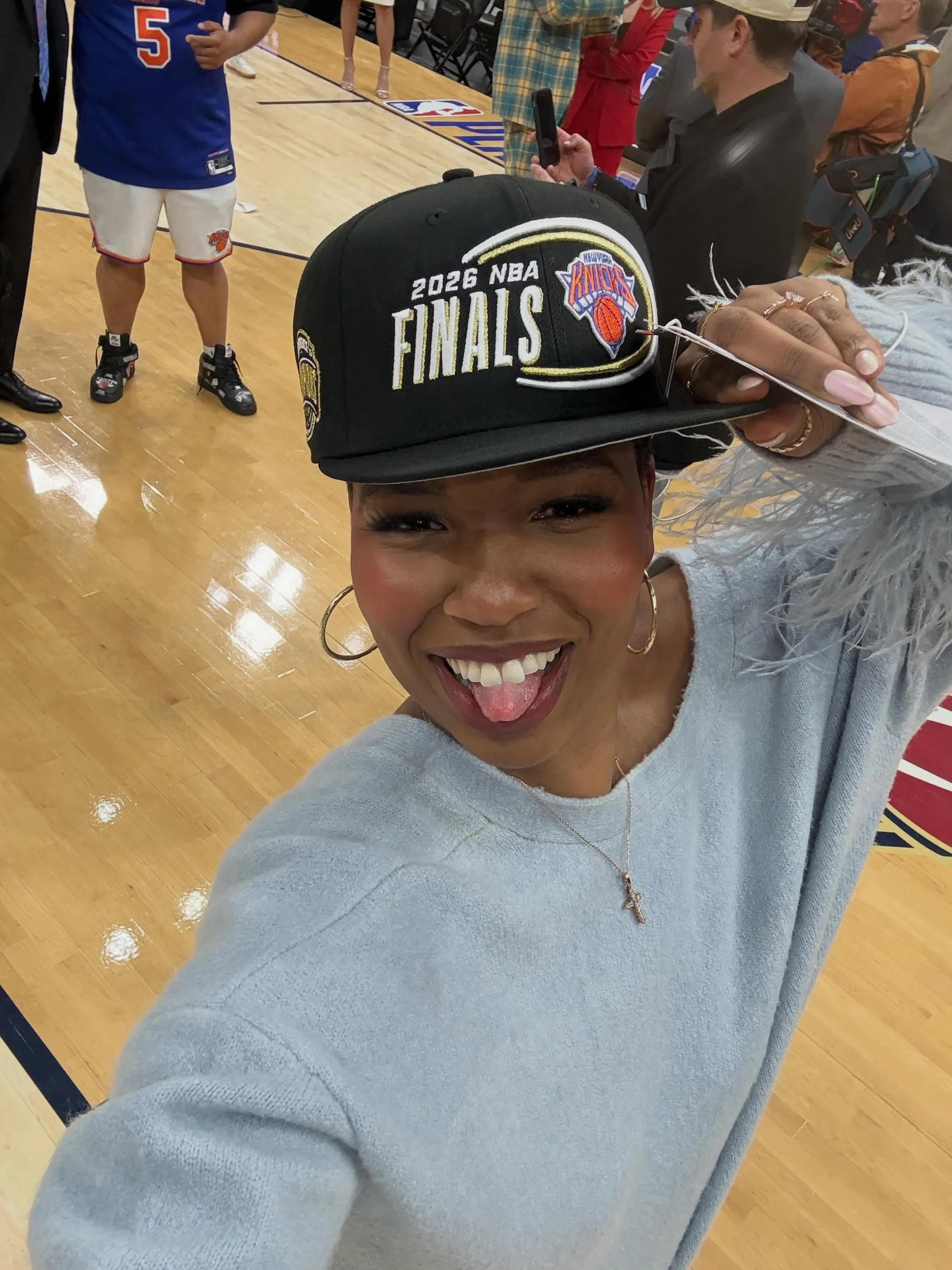 Monica McNutt smiling and holding a black cap that says "2026 NBA FINALS" and has the New York Knicks logo on it.