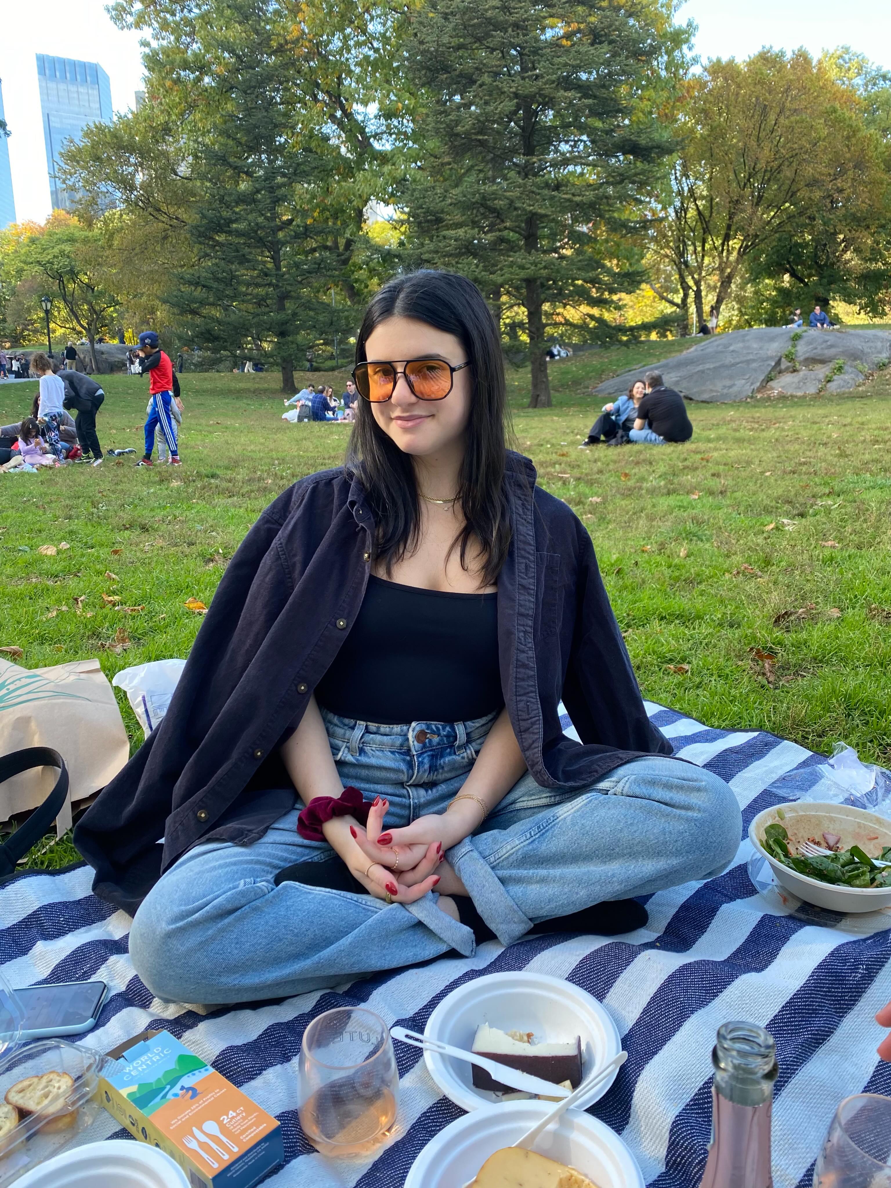 A woman in sunglasses and a black shirt sits cross-legged on a striped blanket with food, drinks, and a phone, smiling at the camera in a park setting with trees, people, and buildings in the background.