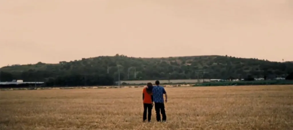 Two people stand in a field with their backs to the viewer, looking at a hill.