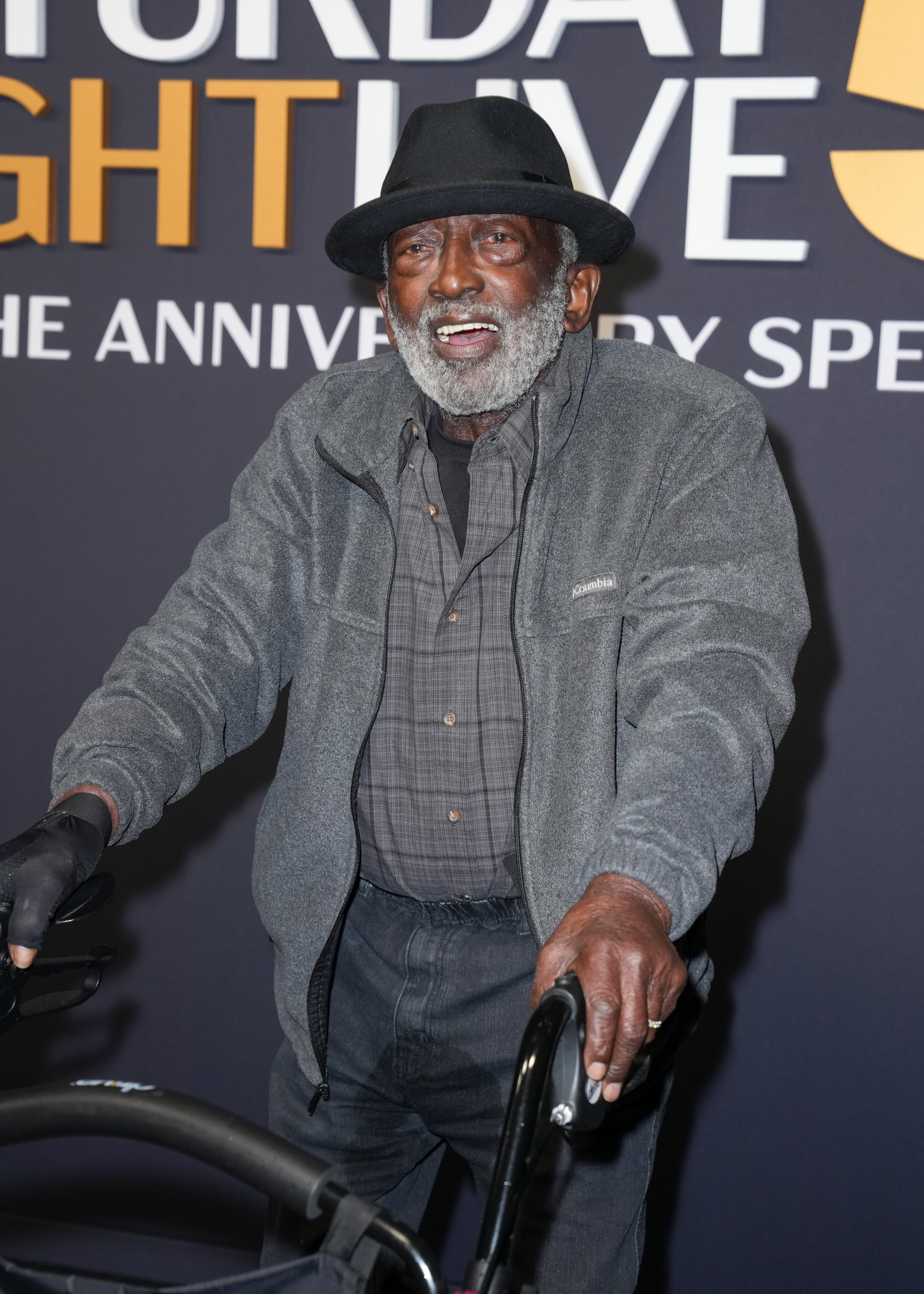 Original "Saturday Night Live" cast member Garrett Morris in a black hat, grey jacket, and plaid shirt, smiling and holding a walker.