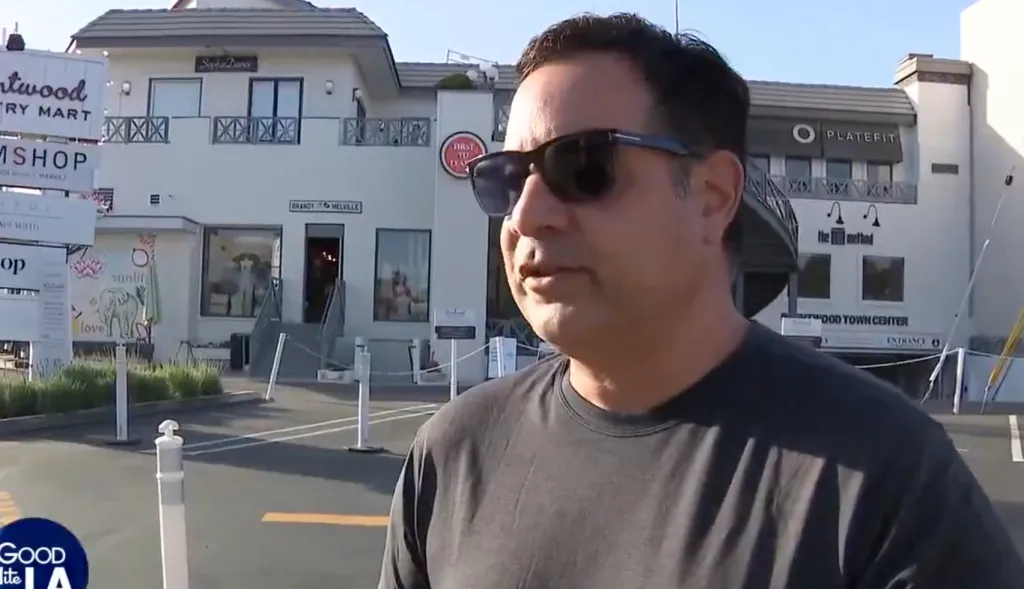 A man in sunglasses being interviewed with "Brentwood Country Mart" signs and shops in the background.