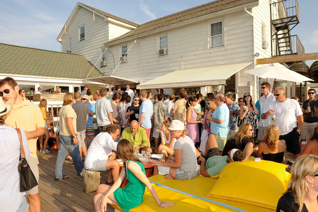 A large group of people socializing outdoors at a party with a two-story white building in the background.