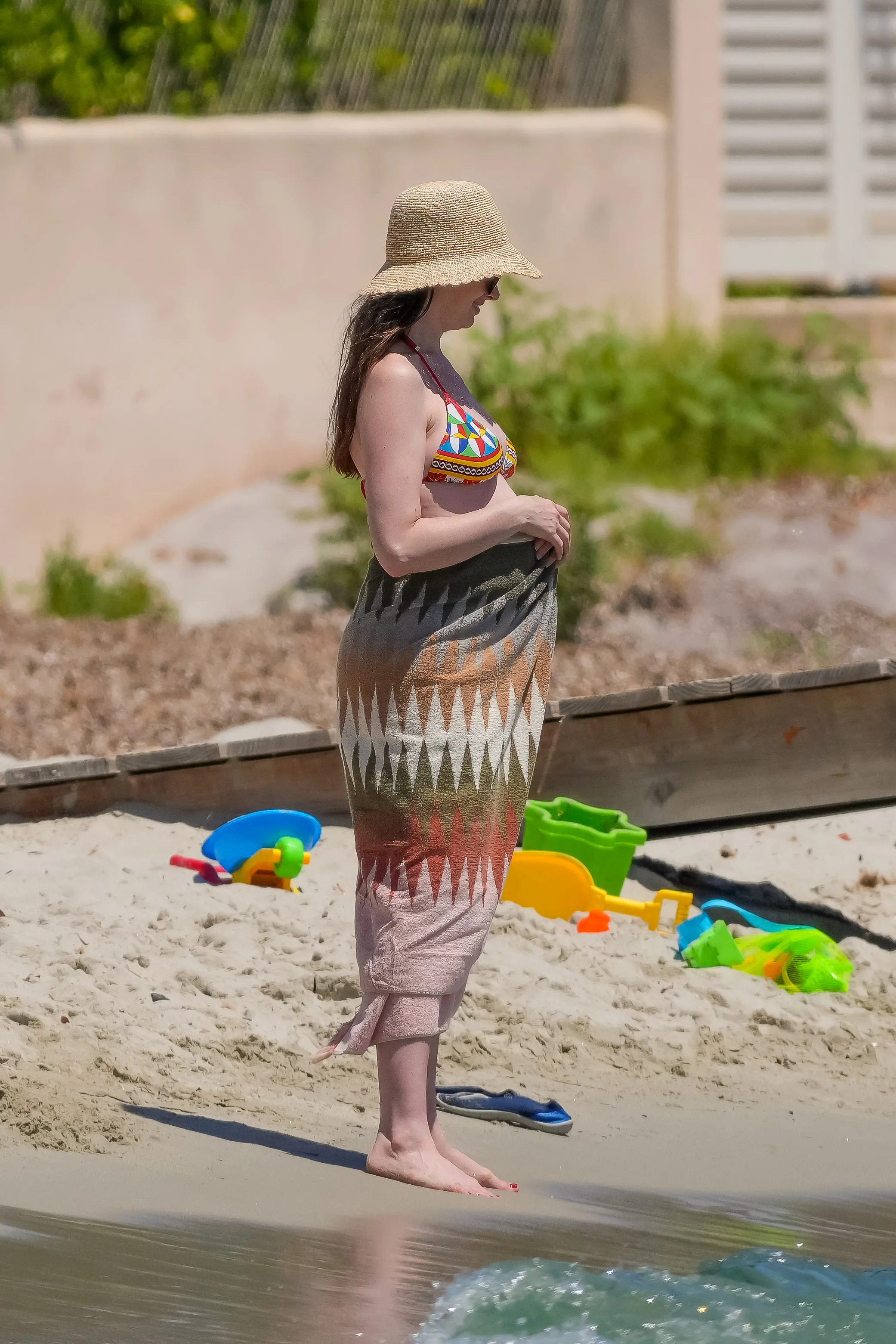 Anne Hathaway in a multi-color bikini top and patterned sarong, standing on a beach.