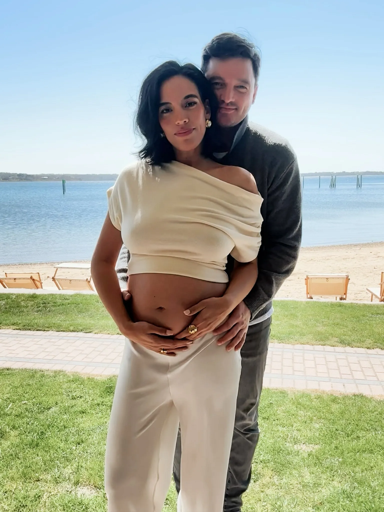 Danielle Olivera and Eoin Heavey posing with her baby bump, with a beach and water in the background.
