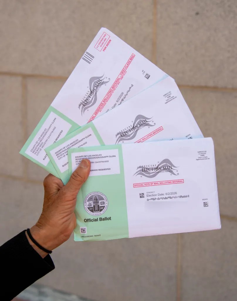 A hand holding several official ballot envelopes marked for election mail, with "Election Date: 6/2/2026" visible on one.