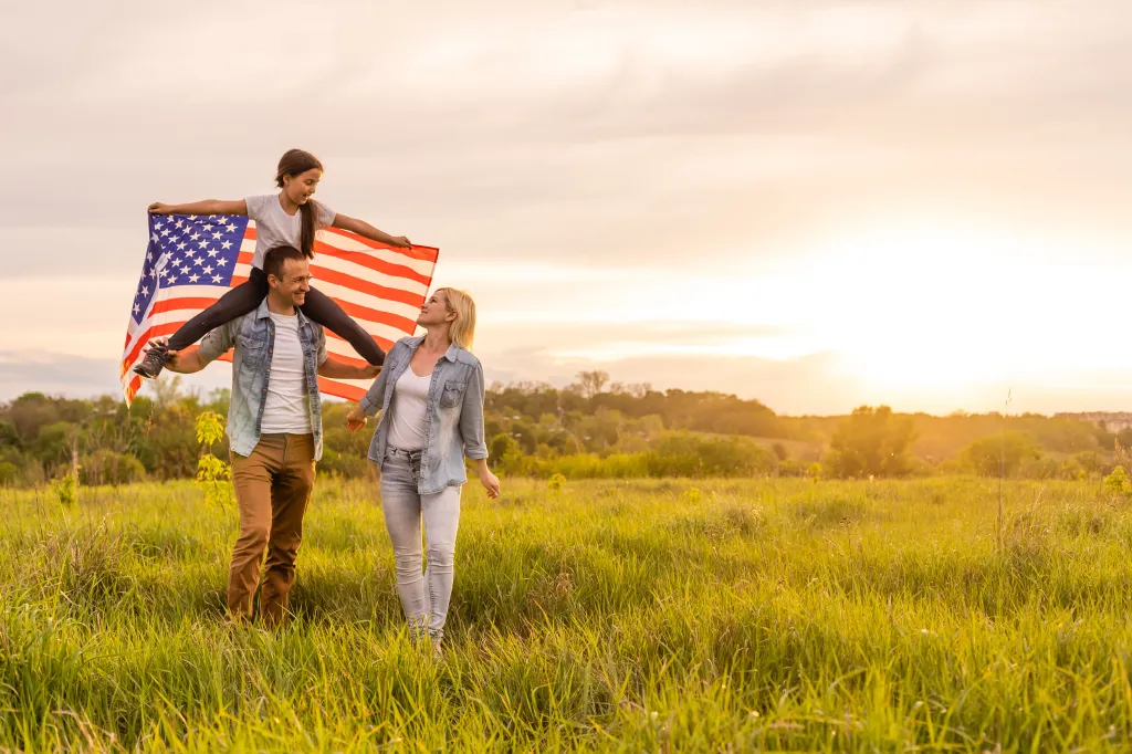 Family with a young girl holding the American flag on her father's shoulders in a field at sunset.