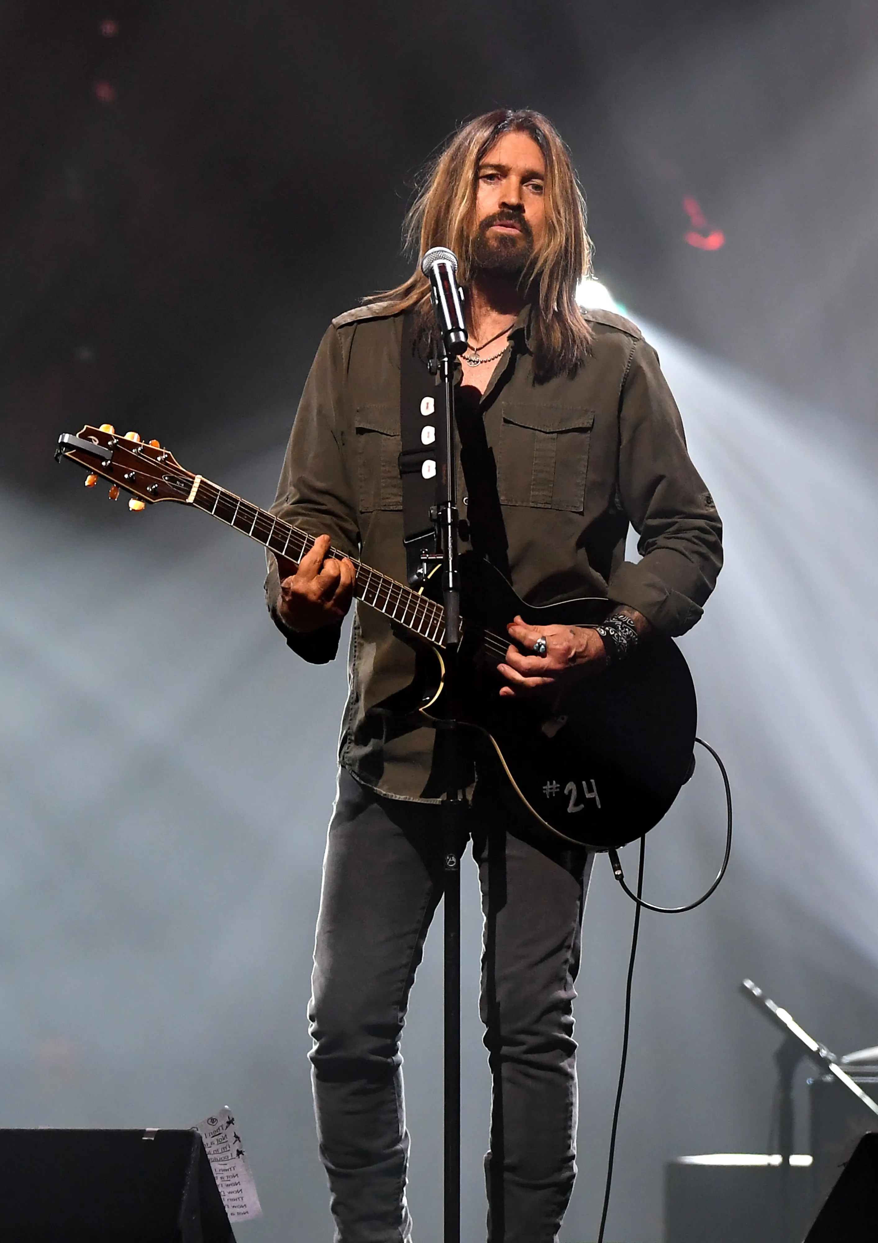Billy Ray Cyrus performing onstage with a black electric guitar.
