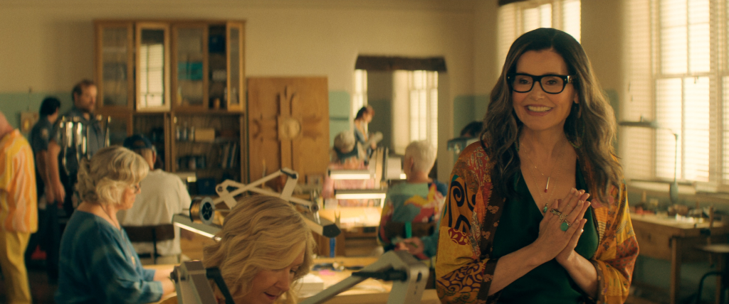 Geena Davis as Renee in "The Boroughs" wearing glasses, a green top, and a colorful patterned robe, smiling with her hands clasped together.