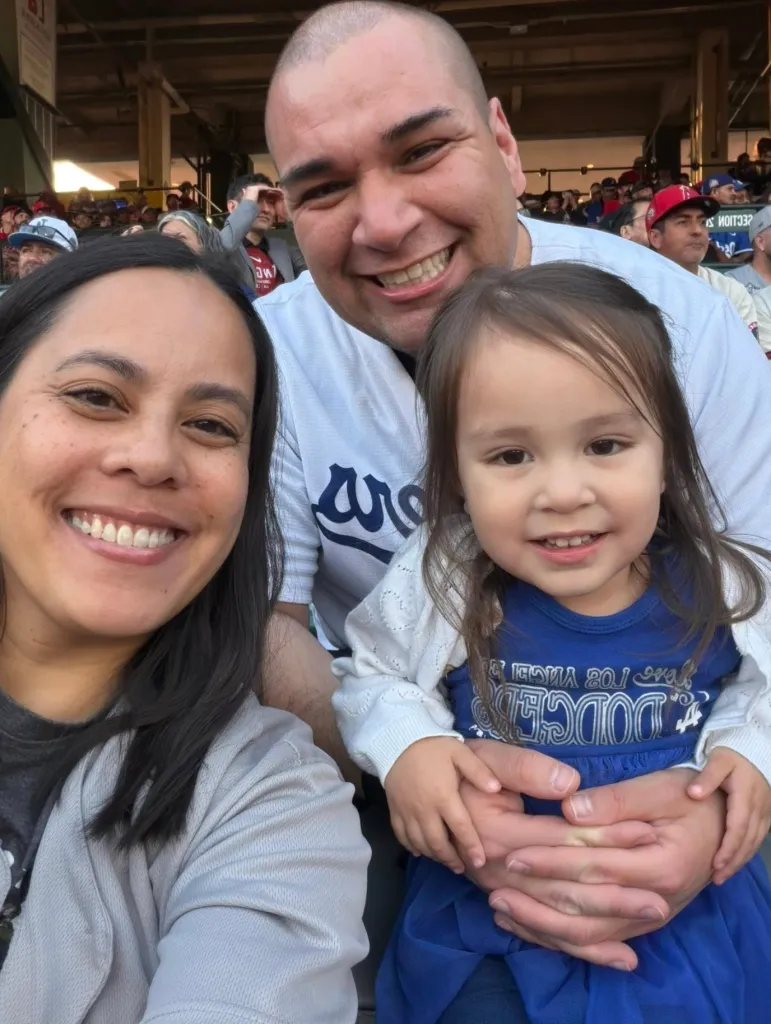 Brianna Nichole, Lt. Colonel Gabriel Estrella, and their daughter smiling at a Dodgers game.