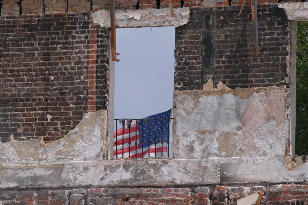 An American flag hangs from a railing in the window of a fire-damaged brick building.