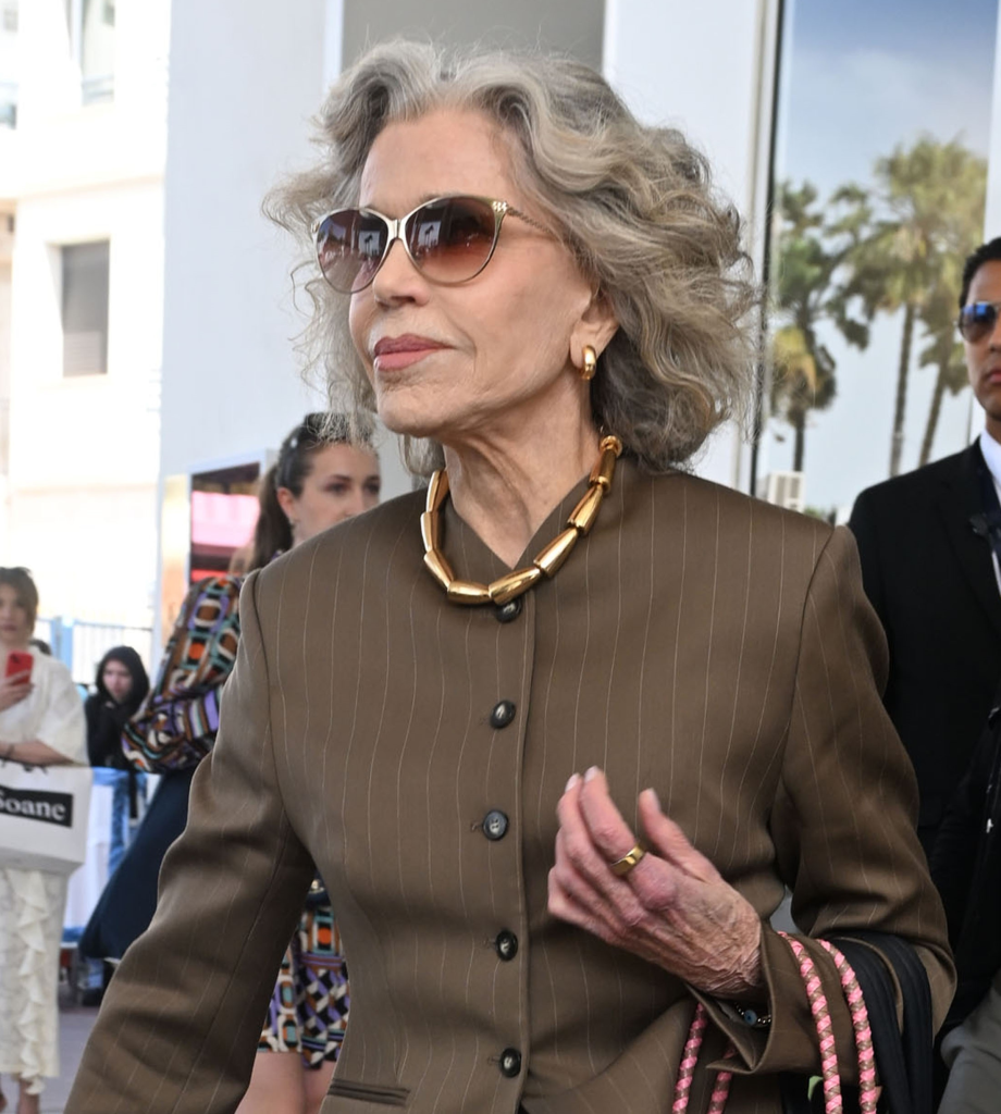 Jane Fonda leaving the Hotel Martinez wearing a tailored brown pinstripe blazer, gold hoop earrings, a chunky link necklace, and oversized gradient sunglasses.