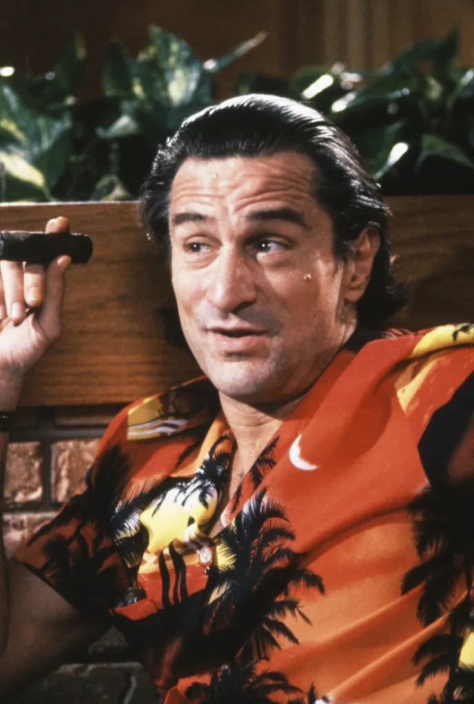 Robert De Niro in a red and yellow tropical print shirt, holding a cigar.