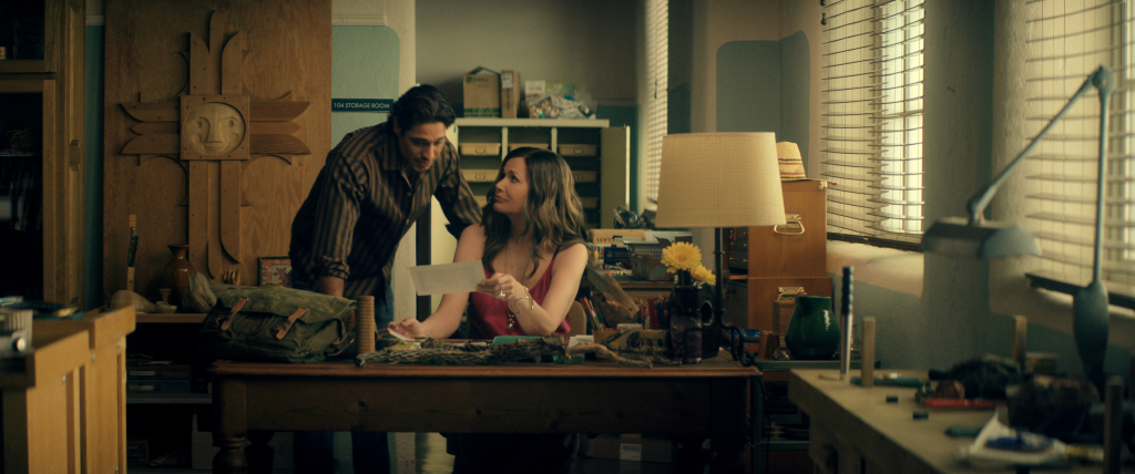 Carlos Miranda as Paz Navarro and Geena Davis as Renee, with Renee seated at a desk holding a document and Paz Navarro standing next to her, in a room with blinds and a wooden sun sculpture on the wall.