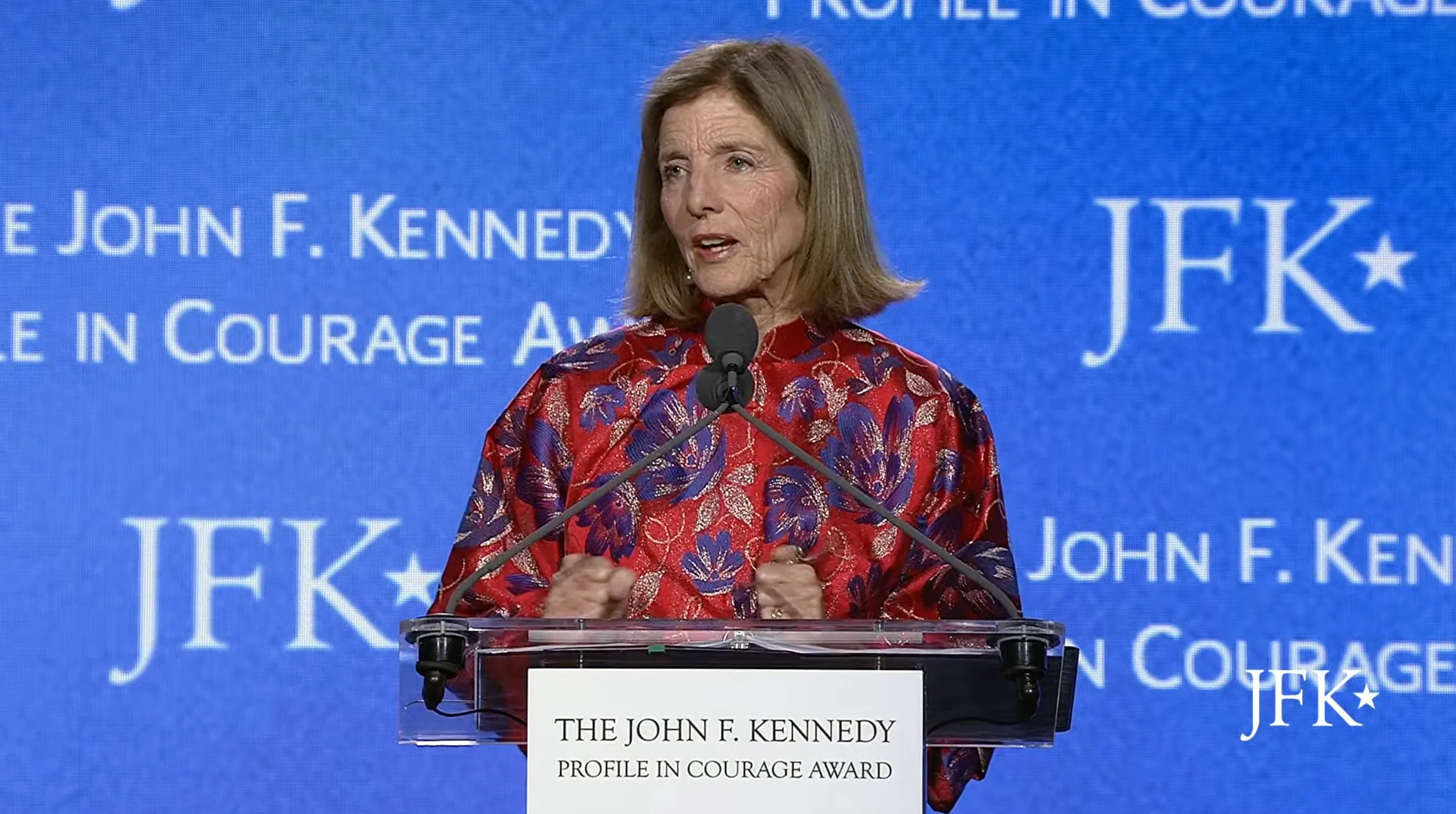 Caroline Kennedy speaking at a podium with "JFK" and "PROFILE IN COURAGE AWARD" visible.
