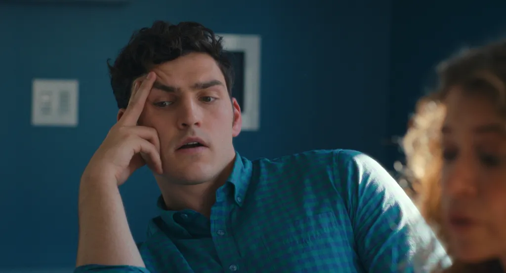 Charlie Hall in a blue and green plaid shirt, looking pensive with his hand to his forehead.