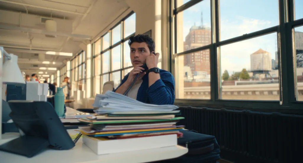 Charlie Hall on the phone at a desk with stacked papers, looking out a large window in "Maximum Pleasure Guaranteed."