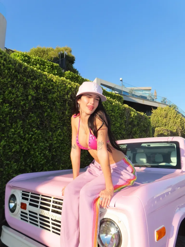 Sami Sheen in a pink bikini top and sweatpants, sitting on a pink custom Ford Bronco.
