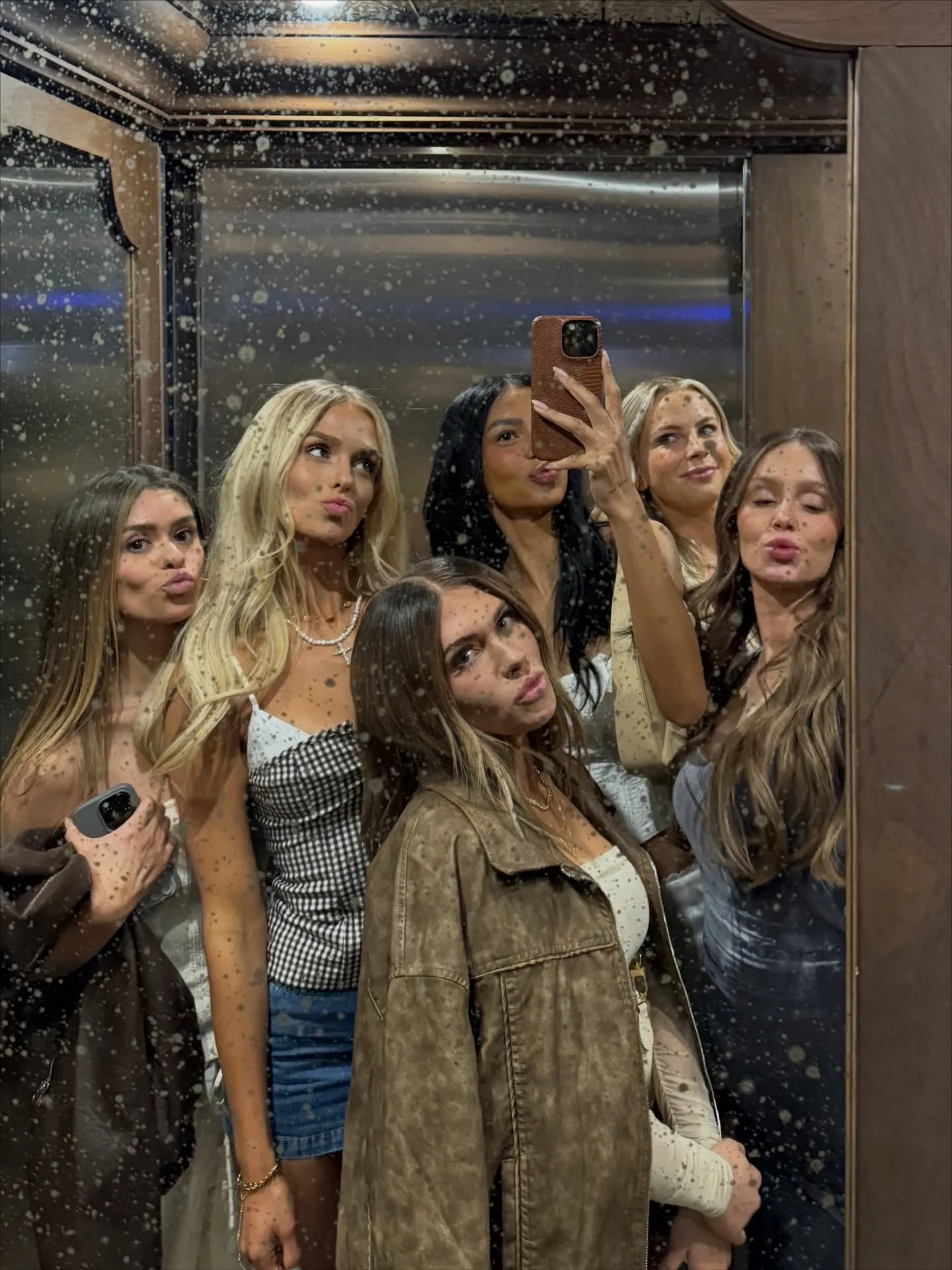Six women taking a selfie in a mirror covered in white speckles.