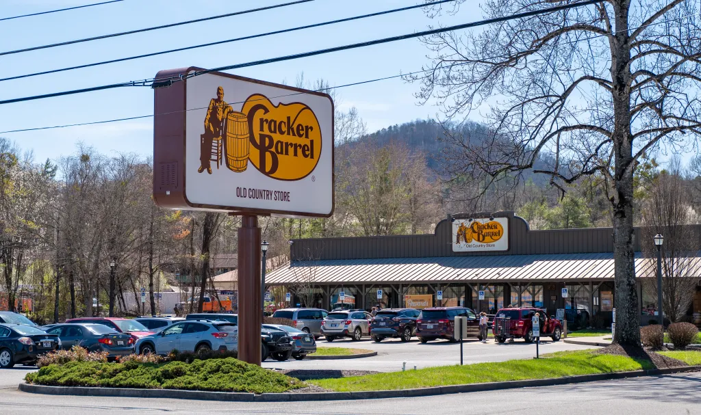 Cracker Barrel Old Country Store sign with a building and busy parking lot in the background.