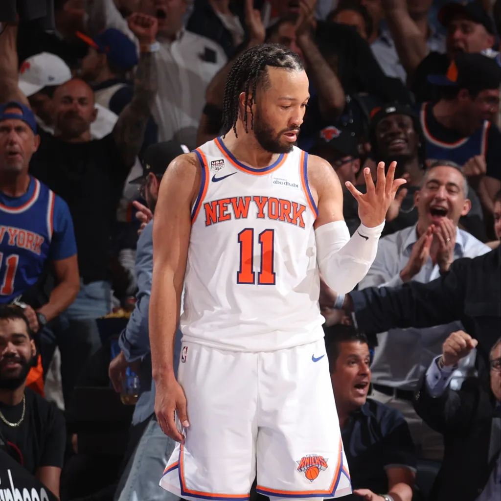 Jalen Brunson in a white New York Knicks jersey, looking down, with fans in the background.
