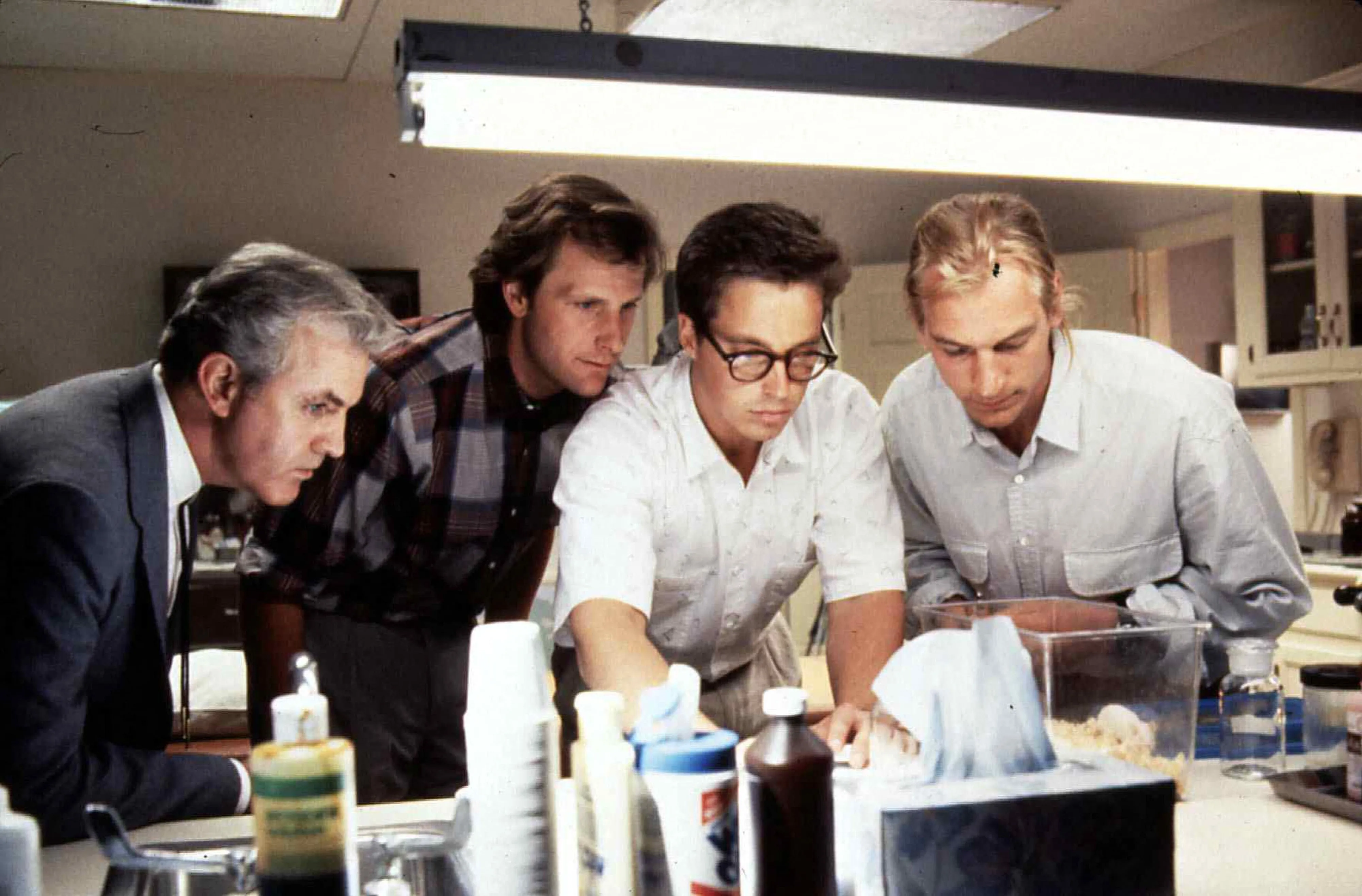 James Handy, Jeff Daniels, Brian McNamara, and Julian Sands leaning over a laboratory counter in a still from the film "Arachnophobia".