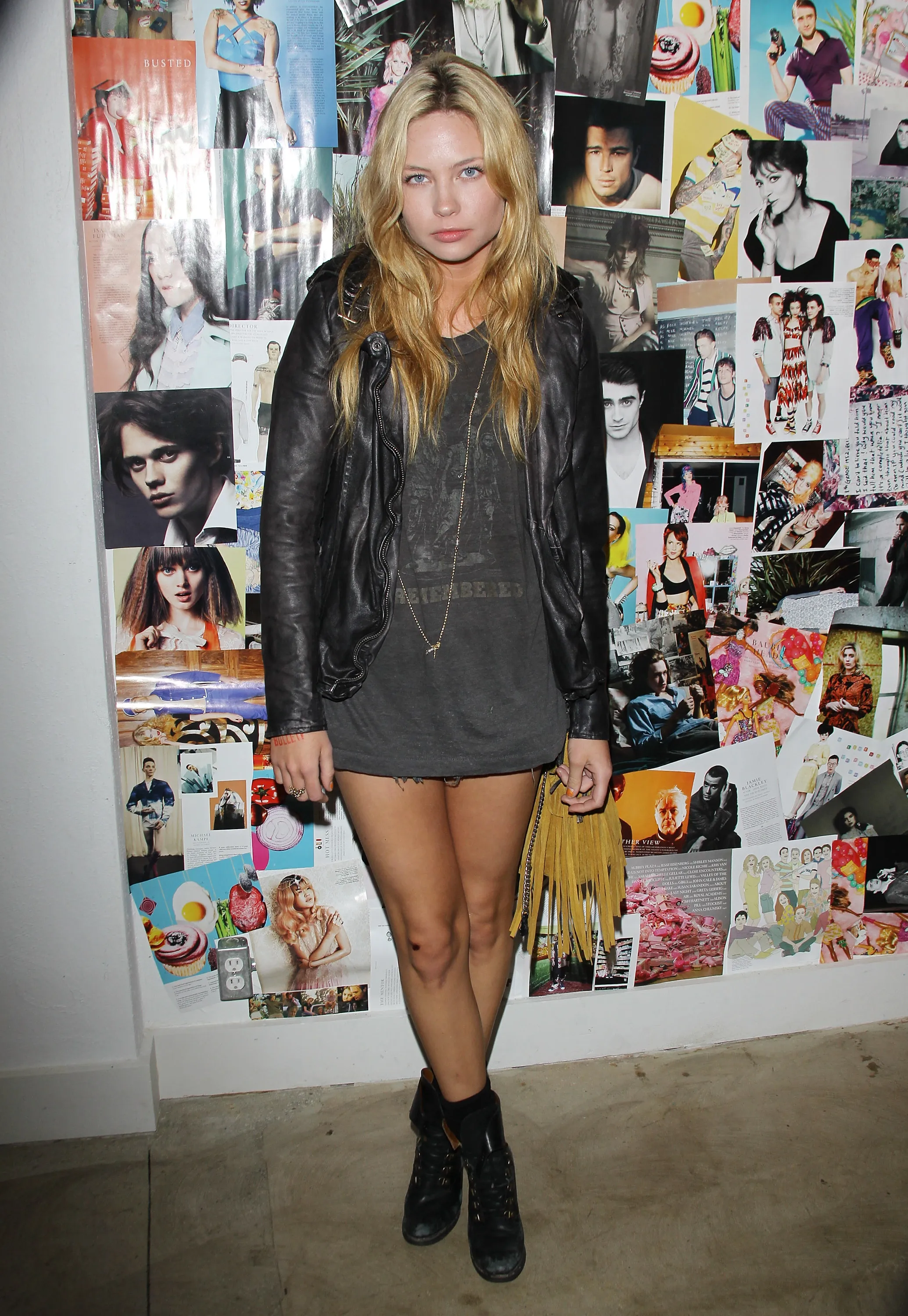 Daveigh Chase wearing a black leather jacket, dark t-shirt, and shorts over dark boots, standing in front of a collage of images.