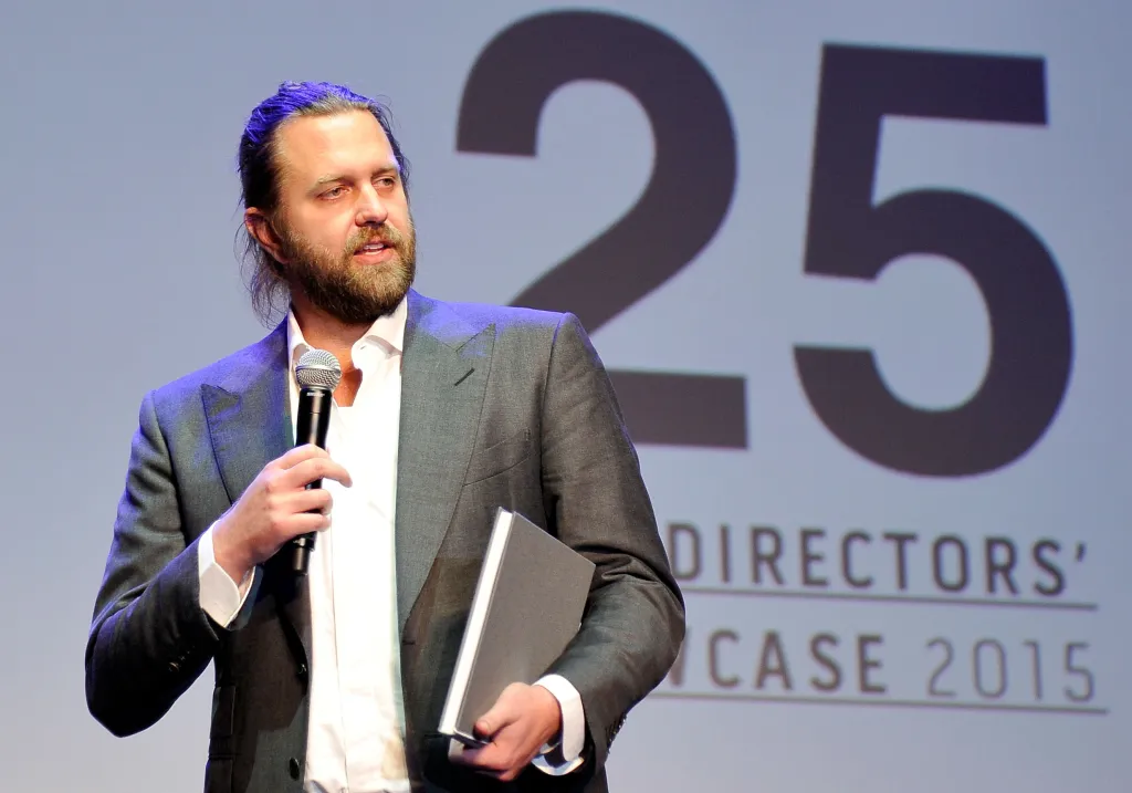Carl Erik Rinsch holding a microphone and a book, standing in front of a screen displaying 