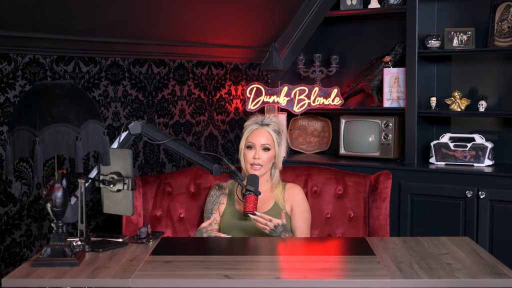 Bunnie XO speaking into a microphone while seated on a red velvet couch in a room with a "Dumb Blonde" neon sign.