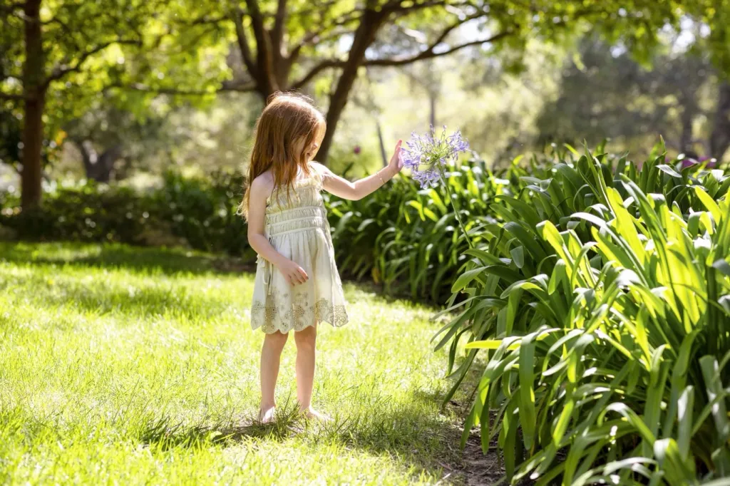 A young girl in a dress touches a purple flower in a sunny garden.