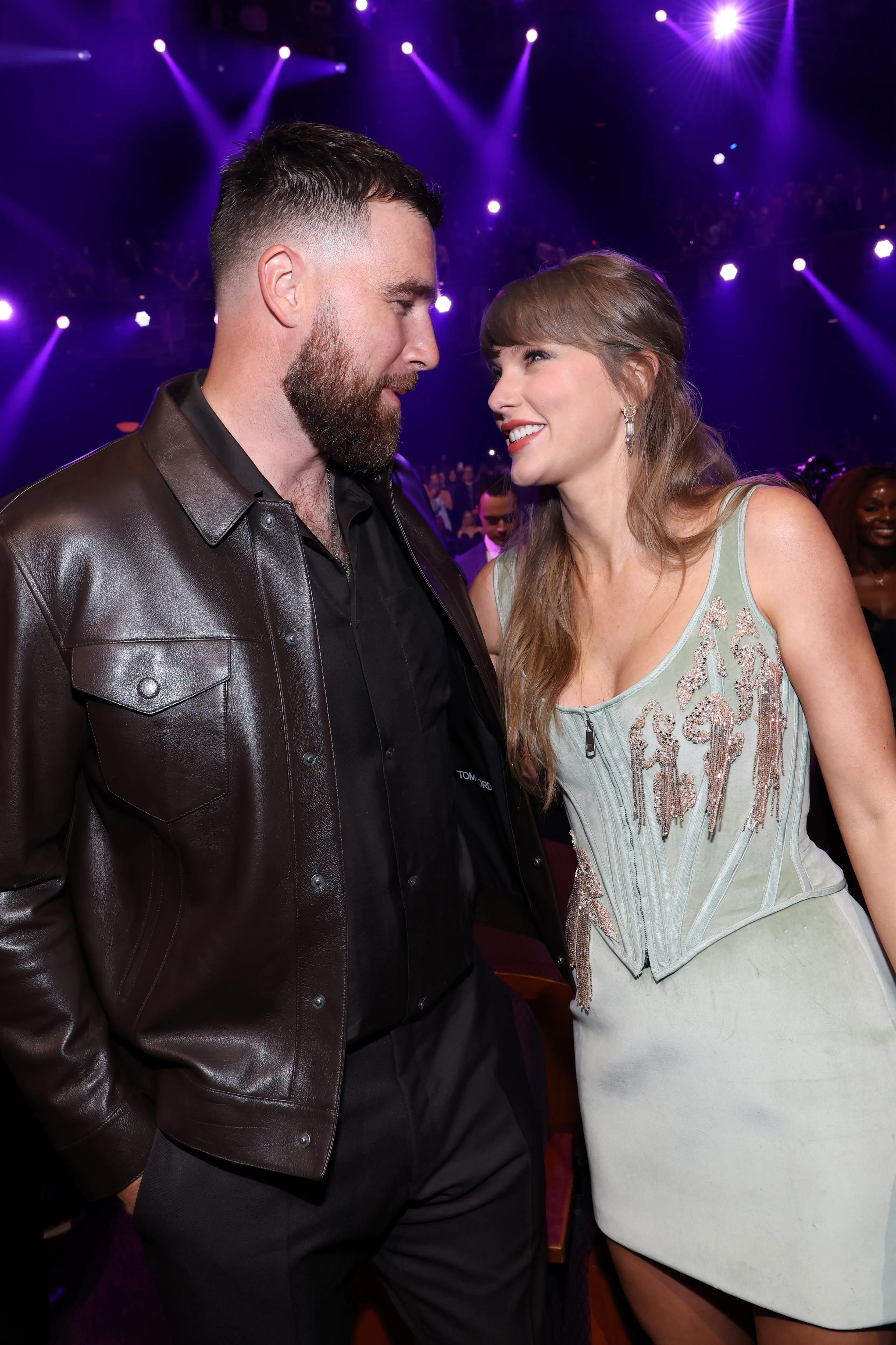 Travis Kelce in a dark brown leather jacket and Taylor Swift in a light blue corset top smiling at each other at the 2026 iHeartRadio Music Awards.